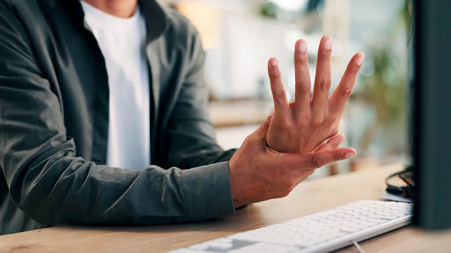 Person holding hand and wrist while sitting at desk with keyboard, representing joint pain affecting daily computer use